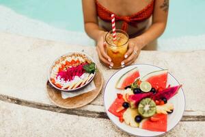 superiore Visualizza su seducente abbronzatura donna nel arancia costumi da bagno rilassante nel piscina con piatto di esotico frutta durante tropicale vacanza. festa umore. foto