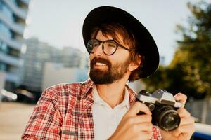 vicino su ritratto di sorridente fricchettone barba uomo utilizzando retrò film telecamera, fabbricazione fotografie mentre in viaggio. urbano sfondo. elegante primavera vestito. indossare nero cappello, scacchi camicia .