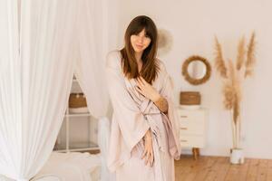 sensuale brunetta donna in posa nel beige seta chimono nel elegante leggero Camera da letto nel boho stile. foto