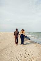 Visualizza a partire dal Indietro. elegante coppia nel amore a piedi lungo il spiaggia. romantico momenti. bianca sabbia e oceano onde. tropicale vacanza. pieno altezza. foto