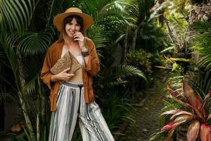 tropicale vacanza e vacanze concetto. elegante elegante ragazza nel bianca superiore e cannuccia cappello in posa su palma le foglie sfondo nel bali. foto