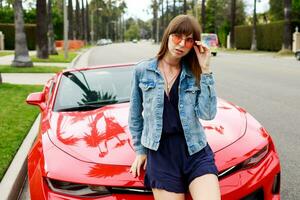 vicino su ritratto di sorpreso brunetta donna seduta su il cappuccio di sorprendente rosso convertibile sport auto nel California. foto