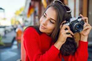 ispirato brunetta donna avendo divertimento e fabbricazione fotografie su sua vacanze . freddo stagione. indossare elegante rosso a maglia maglione. città sfondo.