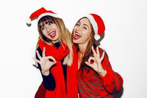 pazzo nuovo anno festa umore. Due ubriaco ridendo donne avendo divertimento e in posa su bianca sfondo nel carino masquerade cappelli. rosso maglione e sciarpa. mostrando ok di mani. foto