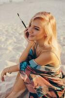 estate stile di vita Immagine di biondo donna disegno acquerello fiore di spazzola. artista seduta su il spiaggia. boemo vestito. foto