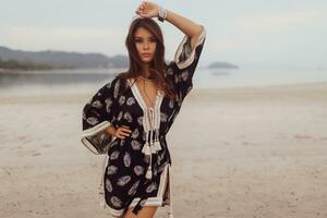 bellissimo asiatico donna nel tropicale boemo vestito in posa su il spiaggia. indossare orecchini con piuma, collana e bracciali. foto