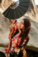 moda foto di elegante asiatico donna nel seta chimono Tenere fan e in posa al di sopra di rocce su il spiaggia.