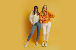 Due bella donne , migliore amici nel elegante autunno casuale Abiti avendo divertimento al di sopra di giallo sfondo nel studio. pieno lunghezza. foto