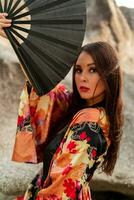 moda foto di elegante asiatico donna nel seta chimono Tenere fan e in posa al di sopra di rocce su il spiaggia.