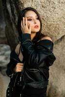 fiducioso donna con rosso labbra nel di moda nero pelle giacca e sensuale nero vestito in posa su roccioso sfondo. foto