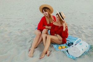 Due bella donne nel rosso estate attrezzatura abd cannuccia cappelli godendo picnic su il spiaggia. estate umore. foto