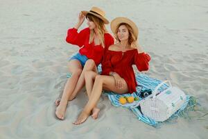 Due bella donne nel rosso estate attrezzatura abd cannuccia cappelli godendo picnic su il spiaggia. estate umore. foto