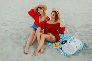 Due bella donne nel rosso estate attrezzatura abd cannuccia cappelli godendo picnic su il spiaggia. estate umore. foto