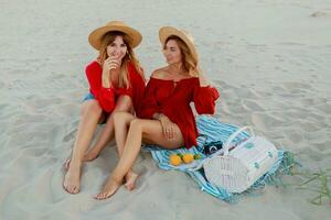 Due bella donne nel rosso estate attrezzatura abd cannuccia cappelli godendo picnic su il spiaggia. estate umore. foto