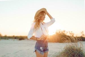 in viaggio e vacanze concetto. contento femmina a piedi su sera spiaggia e Tenere retrò telecamera. indossare cannuccia cappello e sexy jeans corti. caldo tramonto colori . foto