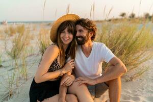 in viaggio coppia nel amore in posa su il spiaggia. brunetta donna nel cannuccia cappello con sua fidanzato chilling nel caldo estate sera. foto