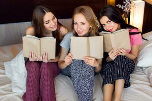 stile di vita Immagine di tre migliore amici nel pigiama lettura libri, studiando e conversazioni nel Camera da letto. gruppo di studenti fare compiti a casa insieme a casa. accogliente caldo colori. emotivo facce. foto