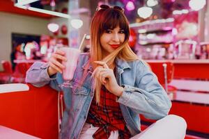 alla moda ragazza nel jeans giacca seduta nel t retrò americano bar. bello sorridente donna godendo dolce latte shake . indossare bianca scarpe da ginnastica. foto