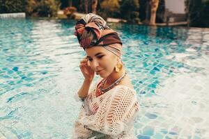 donna con luminosa rendere su nel boemo vestito, turbante, etnico orecchini, collana e bracciali in posa nel nuoto piscina. foto