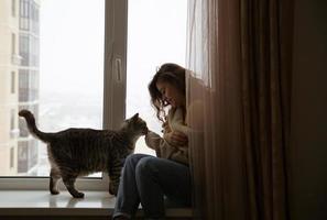 ragazza si siede vicino alla finestra accanto a un gatto foto
