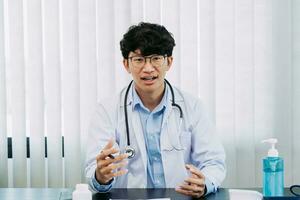 il medico asiatico che indossa occhiali e uniformi bianche con uno stetoscopio fornisce consulenza online ai pazienti durante l'epidemia di virus mantenendo una distanza sociale. foto