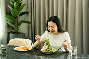 giovane donna asiatica sorridente mentre raccoglie un'insalata su un piatto e mangia felicemente a casa. foto
