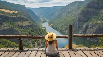 femmina viaggiatore nel cannuccia cappello seduta su di legno pilastro ammirazione mozzafiato scenario di fiume canyon. generativo ai foto