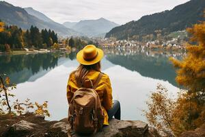 posteriore Visualizza di un' elegante ragazza, con un' zaino, un' cappello e un' giallo giacca, guardare a il Visualizza di il montagne e il lago mentre rilassante nel il autunno natura. viaggio concetto. generativo ai. foto