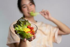 ritratto di un' contento giocoso asiatico ragazza mangiare fresco insalata a partire dal un' bicchiere ciotola dopo allenarsi a casa. giovane signora godendo salutare nutrizione e biologico cibo, avendo vegetariano pasto foto