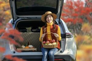 contento asiatico contadino ragazza trasporto produrre raccogliere con homegrown organici mela, schiacciare e zucca seduta su il auto tronco a Locale azienda agricola mercato durante autunno stagione per agricoltura Prodotto foto