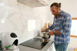 ritratto di un' bello giovane uomo mettendo un' casseruola su il superficie di un elettrico stufa mentre cucinando nel casa cucina foto