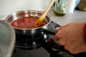 avvicinamento capocuoco mano utilizzando un' di legno cucchiaio, stagionatura e miscelazione un' bollente pomodoro salsa nel un' casseruola su elettrico stufa foto