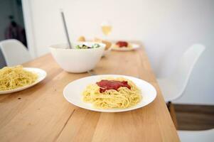 un' di legno tavolo con servito piatti di appena preparato spaghetti con pomodoro salsa nel moderno leggero casa cucina interno foto