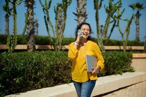 sorridente fiducioso donna, attività commerciale signora parlando su mobile Telefono, Tenere il computer portatile anno Domini passeggiando il strada. le persone. attività commerciale foto