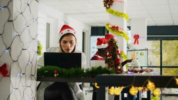 focalizzata dipendente indossare Santa cappello Lavorando su in ritardo progetto durante Natale stagione nel festivo decorato ufficio. diligente personale membro nel diverso posto di lavoro adornano con rosso natale ghirlande e fiocchi foto