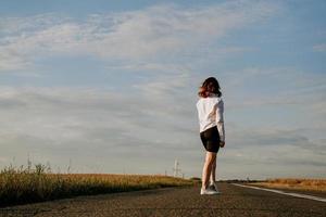 donna in camicia bianca cammina lungo la strada tra i campi foto