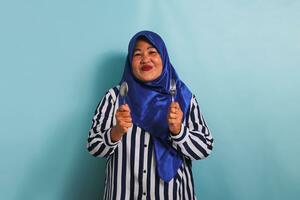un eccitato di mezza età asiatico donna nel un' blu hijab e un' a strisce camicia è Tenere un' cucchiaio e forchetta, sensazione Affamato mentre in attesa per cibo per essere servito. lei è isolato su un' blu sfondo foto