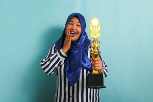 un eccitato di mezza età asiatico donna d'affari nel un' blu hijab e un' a strisce camicia è Tenere un' oro trofeo, festeggiare sua successo e risultato. lei è isolato su un' blu sfondo foto