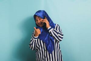 un' allegro di mezza età asiatico donna nel un' blu hijab e un' a strisce camicia è dando un' pollici su mentre parlando su il Telefono. lei è isolato su un' blu sfondo foto