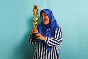 un eccitato di mezza età asiatico donna d'affari nel un' blu hijab e un' a strisce camicia è Tenere un' oro trofeo, festeggiare sua successo e risultato. lei è isolato su un' blu sfondo foto