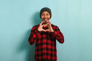 sorridente giovane asiatico uomo Spettacoli un' amore gesto con il suo mano mentre in piedi contro un' blu sfondo foto