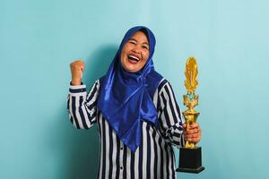 un' orgoglioso di mezza età asiatico donna d'affari nel un' blu hijab e un' a strisce camicia è fabbricazione un' vincitore gesto mentre Tenere un' oro trofeo, festeggiare sua successo. lei è isolato su un' blu sfondo. foto