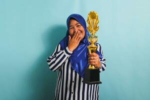 un eccitato di mezza età asiatico donna d'affari nel un' blu hijab e un' a strisce camicia è Tenere un' oro trofeo, festeggiare sua successo e risultato. lei è isolato su un' blu sfondo foto