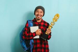 contento giovane asiatico uomo, Tenere un' oro trofeo, gioia nel il suo successo, isolato su blu sfondo. foto