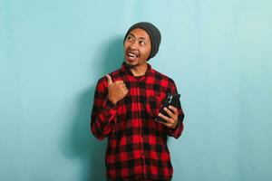 giovane asiatico uomo con un' Beanie cappello e un' rosso plaid flanella camicia è puntamento il suo dito sinistra a copia spazio mentre Tenere un' mobile Telefono, isolato su un' blu sfondo foto