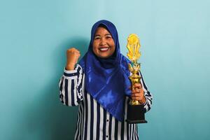 un' orgoglioso di mezza età asiatico donna d'affari nel un' blu hijab e un' a strisce camicia è fabbricazione un' vincitore gesto mentre Tenere un' oro trofeo, festeggiare sua successo. lei è isolato su un' blu sfondo. foto