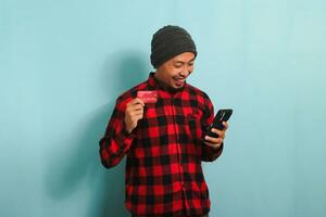 contento giovane asiatico uomo con un' Beanie cappello e un' rosso plaid flanella camicia è Tenere un' smartphone e un' bancario credito carta, fare in linea acquisti, isolato su un' blu sfondo. foto