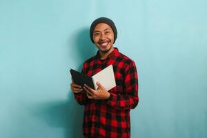 un' sorridente giovane asiatico uomo con un' Beanie cappello e un' rosso plaid flanella camicia è Tenere un' libro mentre in piedi contro un' blu sfondo foto