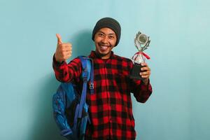 eccitato giovane asiatico uomo alunno dando pollice su mentre Tenere un' trofeo, isolato su blu sfondo foto