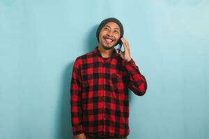 contento giovane asiatico uomo con Beanie cappello e rosso plaid flanella camicia è ridendo mentre parlando su il suo smartphone, avendo un' bene conversazione, udito divertente scherzi, isolato su un' blu sfondo foto